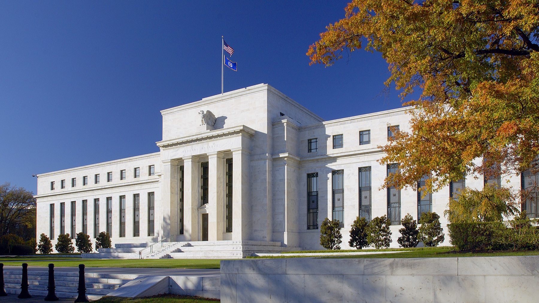 Three-quarter exterior view of the white marble Marriner S. Eccles Building under a deep blue sky, with autumn foliage to the right.