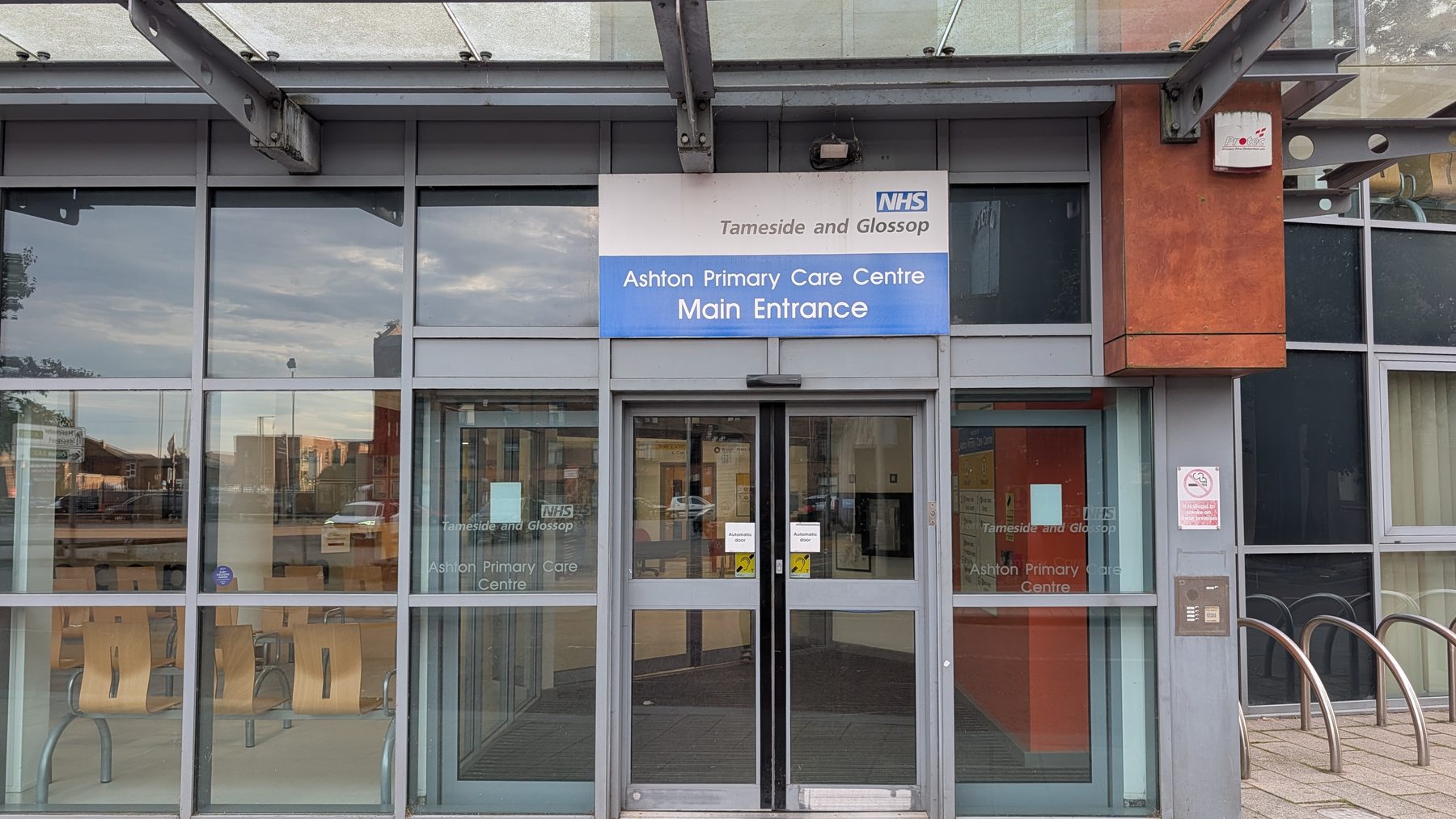 Glazed automatic doors beneath a steel canopy at the main entrance of Ashton Primary Care Centre, with an NHS Tameside and Glossop sign above.