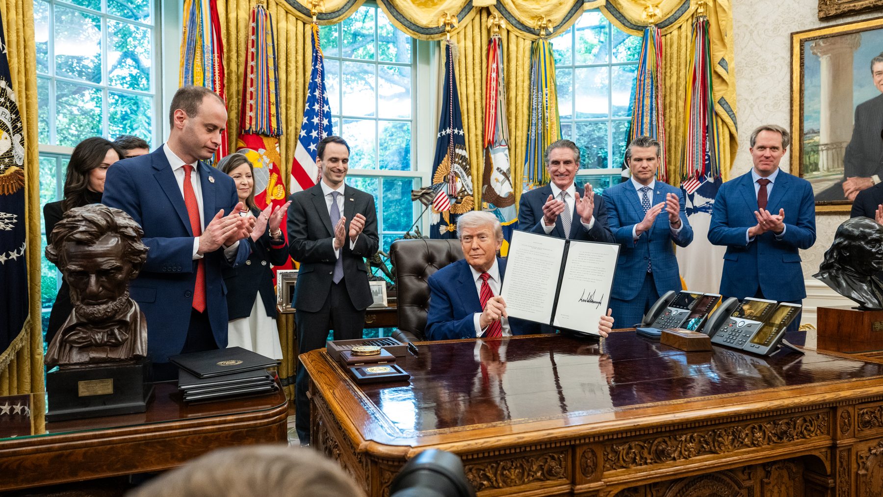 President Donald Trump seated at the Oval Office desk holding up a signed executive order, surrounded by applauding officials.